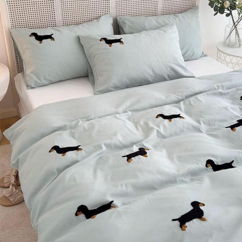 May include: A light blue bedding set featuring embroidered black dachshund dogs. The set includes a duvet cover and pillowcases. The bed is made up with a white mattress and a woven headboard.