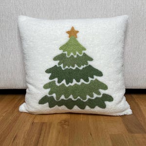 May include: White square pillow with a green Christmas tree design. The tree is made of various shades of green and topped with a gold star. The pillow has a soft, textured appearance, perfect for holiday decor.