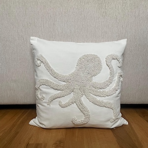 Op de afbeelding: Vierkant wit kussen met een lichtgrijs octopusontwerp. De octopus is geborduurd op het kussen. Het kussen ligt op een houten vloer. Een decoratief accent voor een kust- of nautisch thema, ongeveer 40 cm x 40 cm.