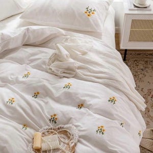 May include: White bedding set with embroidered yellow floral details. The set includes a duvet cover and pillowcases. A decorative tray with a bar of soap and a string of pearls is on the bed.
