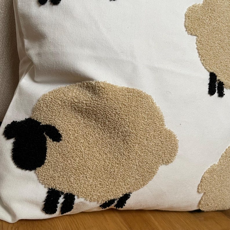 May include: White throw pillow with a repeating sheep design. The sheep have tan wool-like bodies, black heads and legs. The pillow is made of a soft, textured fabric, perfect for a farmhouse or country-themed decor.