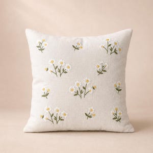 May include: A square throw pillow in a light beige fabric featuring scattered embroidered daisy motifs. The flowers have white petals and yellow centers with green stems and leaves, arranged in a repeating pattern across the front surface.