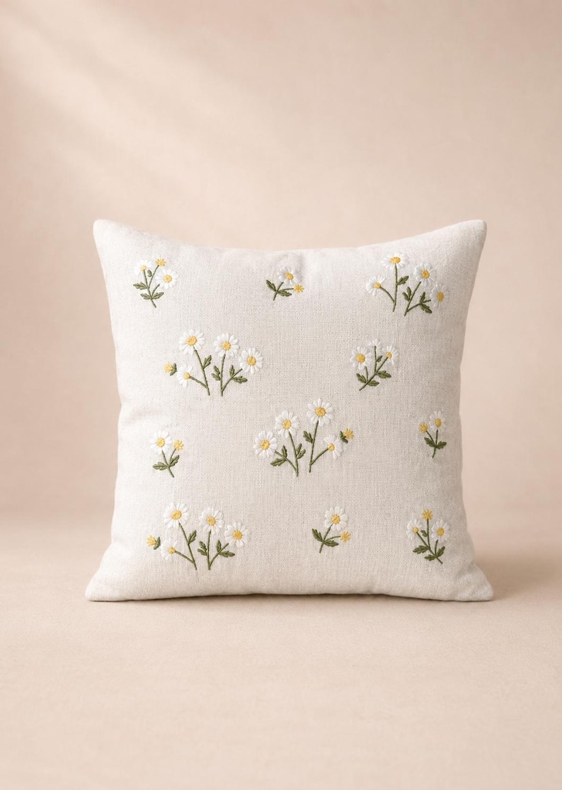 May include: A square throw pillow in a light beige fabric featuring scattered embroidered daisy motifs. The flowers have white petals and yellow centers with green stems and leaves, arranged in a repeating pattern across the front surface.