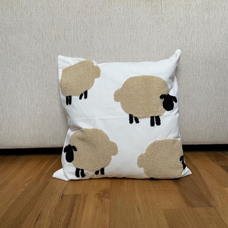 May include: White square pillow with four beige sheep designs. Each sheep has a textured, fluffy appearance with black legs and a black face. The pillow is placed on a wooden floor.