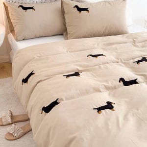 May include: Beige bedding set featuring embroidered black dachshund dogs. The set includes a comforter and two pillowcases. A pair of beige sandals with white ruffles are on the floor. The bedding is on a white bed.