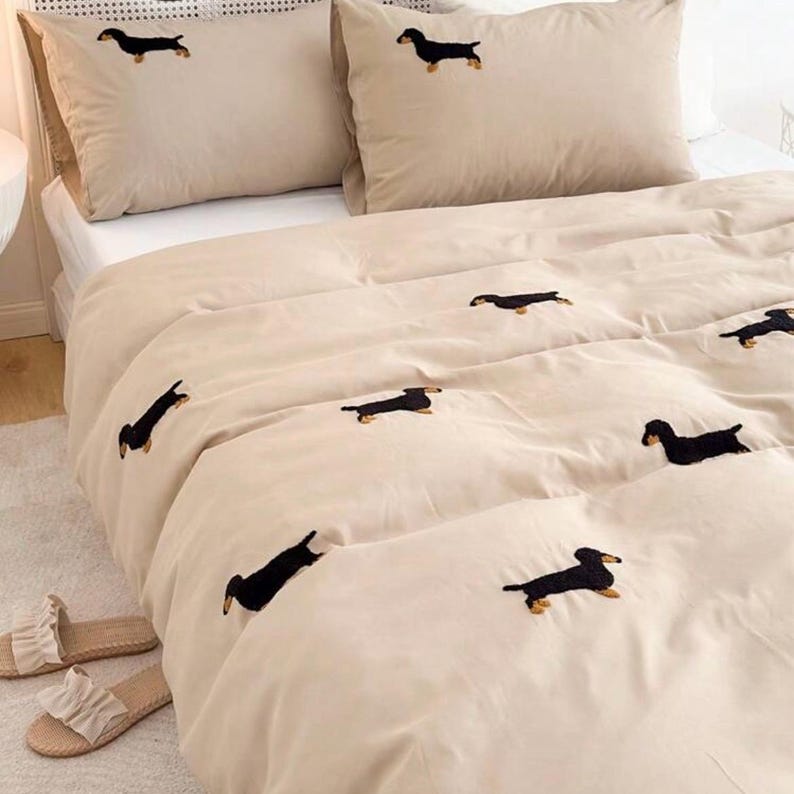 May include: Beige bedding set featuring embroidered black dachshund dogs. The set includes a comforter and two pillowcases. A pair of beige sandals with white ruffles are on the floor. The bedding is on a white bed.