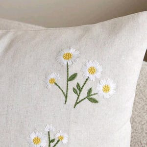 May include: A light beige throw pillow cover featuring embroidered daisy flowers with white petals and yellow centers, accompanied by green stems and leaves. The fabric has a visible woven texture, and the floral design is repeated across the surface.