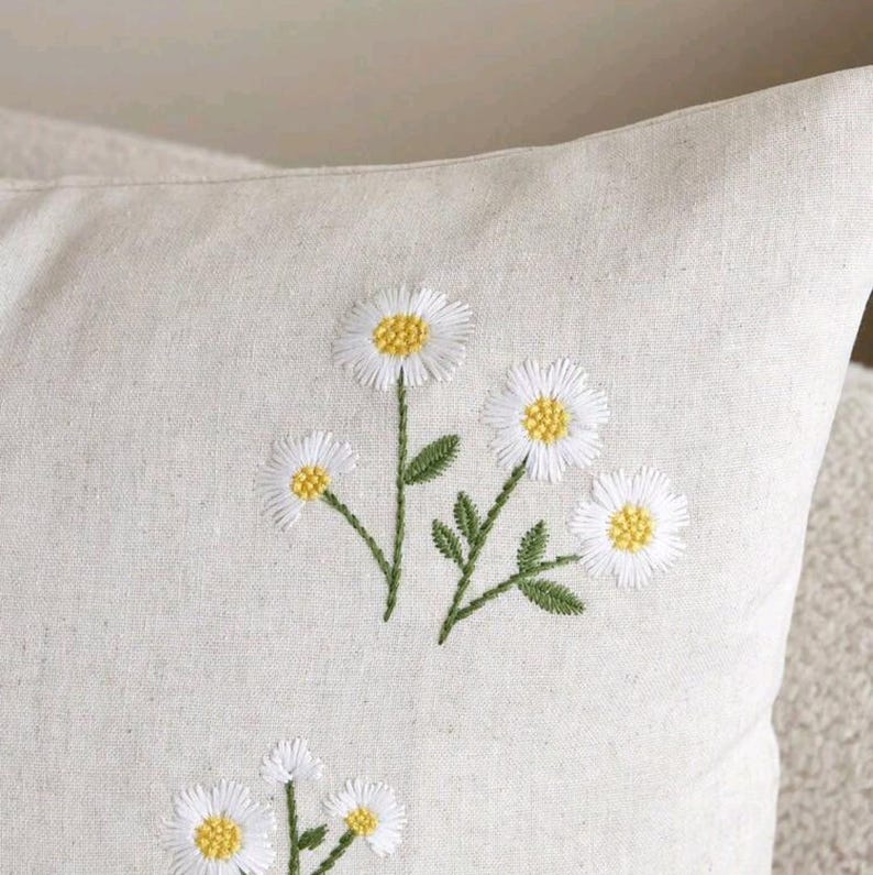 May include: A light beige throw pillow cover featuring embroidered daisy flowers with white petals and yellow centers, accompanied by green stems and leaves. The fabric has a visible woven texture, and the floral design is repeated across the surface.