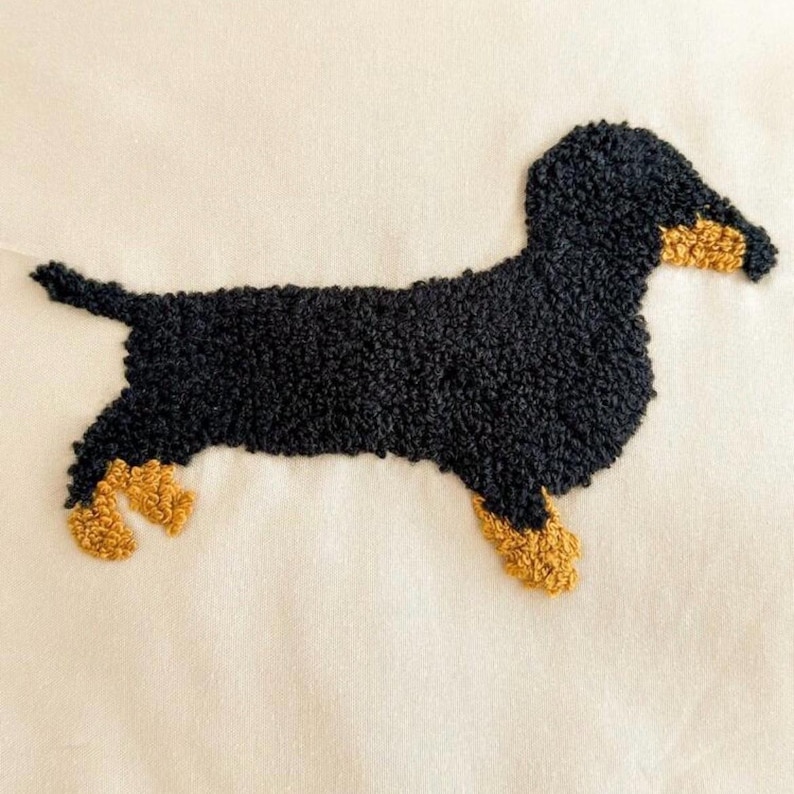 May include: A black and tan dachshund dog design, created with a textured, tufted technique. The dog is depicted in profile, with its body in black and its paws and snout in tan. The background is a cream color.