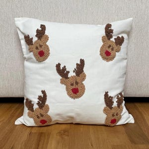 May include: White square pillow with five embroidered reindeer faces. Each reindeer has brown antlers, a tan face, and a red nose. The pillow is placed on a wooden surface.