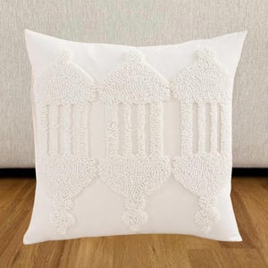 Könnte beinhalten: Ein quadratisches, weißes Zierkissen mit einem strukturierten, erhabenen Design. Das Design zeigt drei stilisierte Laternenformen in einem sich wiederholenden Muster. Das Kissen ist aus einem weichen Stoff gefertigt und eignet sich für ein Bett oder Sofa.