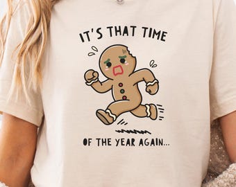 May include: Beige t-shirt featuring the text "IT'S THAT TIME OF THE YEAR AGAIN..." and a cartoon gingerbread man running. The gingerbread man is brown with black details and a worried expression.