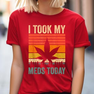 Könnte beinhalten: Rotes T-Shirt mit Retro-Design, das eine Cannabisblatt-Silhouette in Orange-, Gelb- und Grüntönen vor einem Sonnenuntergangshintergrund zeigt. Der Text "I TOOK MY MEDS TODAY" ist oberhalb und unterhalb der Grafik gedruckt.