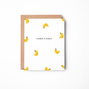 May include: A white greeting card featuring a repeating pattern of yellow banana bunches and the text THANKS A BUNCH printed in black in the centre, accompanied by a brown kraft paper envelope positioned behind the card.