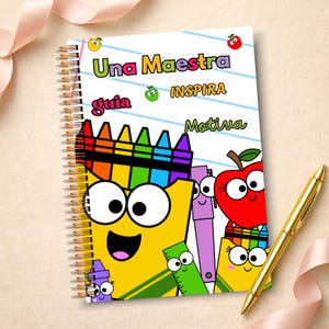 May include: A spiral-bound notebook with a white cover featuring colorful cartoon illustrations of a yellow crayon, a red apple, a purple pen, and a green ruler. The text reads Una Maestra INSPIRA guia Motiva. A gold pen lies next to the notebook.