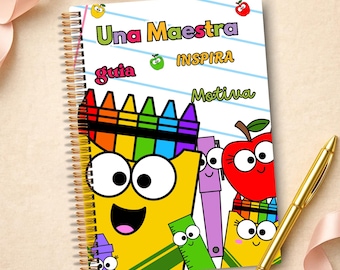 May include: A spiral-bound notebook with a white cover featuring colorful cartoon illustrations of a yellow crayon, a red apple, a purple pen, and a green ruler. The text reads Una Maestra INSPIRA guia Motiva. A gold pen lies next to the notebook.