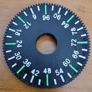 Puede incluir: Un dial circular negro con números y marcas blancas, y detalles en verde. El dial tiene un agujero central y un borde exterior dentado. Los números del 6 al 96 se muestran alrededor del perímetro.