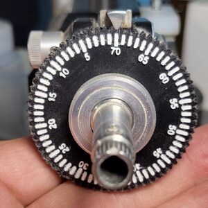 May include: Close-up of a black and silver dial with white markings from 5 to 70. The dial is part of a larger machine, with a silver cylindrical component in the center. The dial is held in a person's hand.