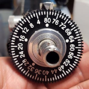 Puede incluir: Primer plano de un dial negro y plateado con marcas blancas. El dial tiene números del 4 al 80, con un cilindro central plateado. El dial es parte de un dispositivo mecánico.