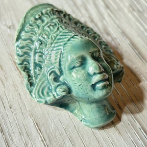 Könnte beinhalten: Eine türkisfarbene Keramikskulptur eines ruhigen Gesichts mit detaillierten Haar- und Gesichtszügen. Die Skulptur ist eine Büste, bei der Kopf und oberer Hals sichtbar sind. Das Stück hat eine glänzende Oberfläche und ist vor einem hellen, strukturierten Hintergrund platziert.