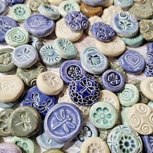 Puede incluir: Una colección de pequeñas baldosas decorativas de cerámica en varios tonos de azul, verde y crema. Las baldosas presentan diseños en relieve como libélulas, flores y patrones abstractos. Las baldosas son redondas y ovaladas, con un acabado brillante.