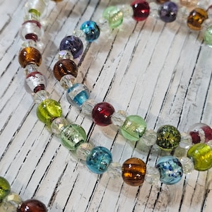 Puede incluir: Un collar de cuentas de vidrio multicolores con cuentas transparentes, marrones, verdes, azules, rojas y moradas. El collar está sobre una superficie de madera blanca.