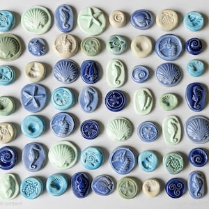 Può includere: Una collezione di piccoli cabochon in ceramica rotondi e ovali in varie tonalità di blu, verde e crema. Ogni pezzo presenta disegni nautici in rilievo tra cui conchiglie, cavallucci marini, stelle marine, tartarughe e motivi a spirale.