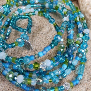 May include: A beaded necklace and matching ring set in shades of blue, green, and clear. The necklace features various bead shapes and sizes, creating a layered effect. The jewelry is displayed on a sandy surface.