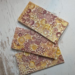 May include: Three rectangular tiles featuring an intricate, embossed floral and paisley pattern in shades of yellow, orange, and deep red. The surface of each tile is covered in a dense, repeating arrangement of various flower shapes, swirls, and leaf motifs.