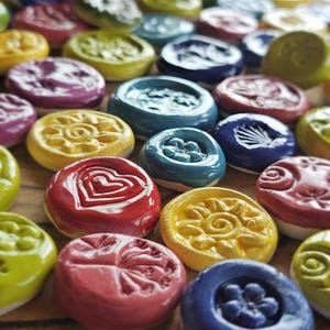 Puede incluir: Una colección de objetos redondos de cerámica colorida con varios diseños en relieve, incluyendo corazones, flores y soles. Los objetos son en tonos de rojo, amarillo, azul, verde y morado. Están dispuestos sobre una superficie de madera.