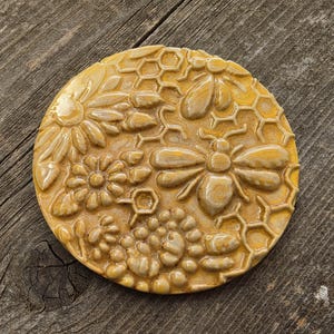 May include: A round, yellow ceramic coaster with a raised relief design featuring bees, flowers, and honeycomb patterns. The coaster is a warm, golden color and appears to be handmade.