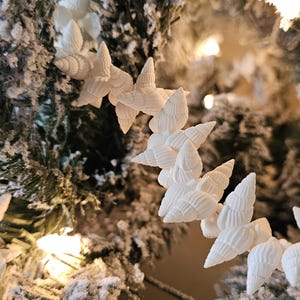 K&ouml;nnte beinhalten: Wei&szlig;e Muschelschmuckst&uuml;cke, die an einem schneebedeckten k&uuml;nstlichen Weihnachtsbaumzweig befestigt sind.