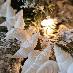 K&ouml;nnte beinhalten: Wei&szlig;e Muscheln, die als Girlande an einem verschneiten k&uuml;nstlichen Weihnachtsbaum aufgeh&auml;ngt sind.