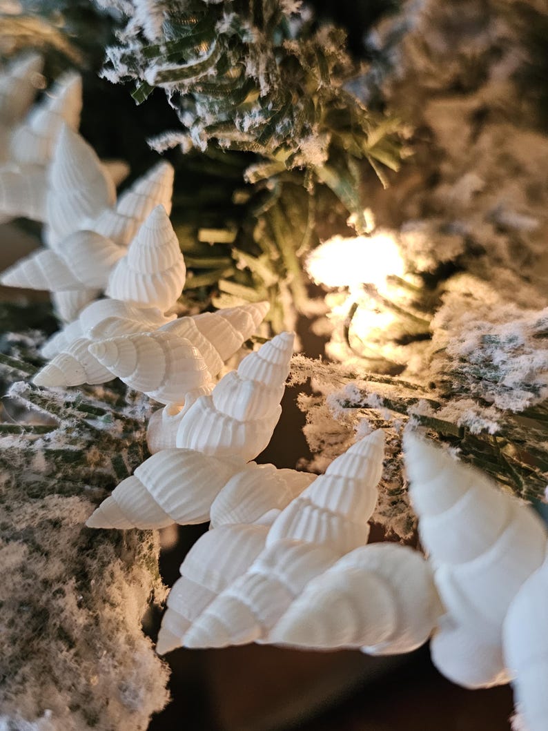 K&ouml;nnte beinhalten: Wei&szlig;e Muscheln, die als Girlande an einem verschneiten k&uuml;nstlichen Weihnachtsbaum aufgeh&auml;ngt sind.