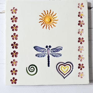 May include: A white ceramic tile with a sun, dragonfly, heart, and spiral design. The tile is decorated with a border of small pink flowers.