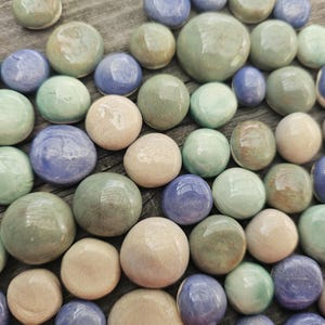 Può includere: Una collezione di perline rotonde in ceramica liscia in vari colori, tra cui verde chiaro, blu e beige. Le perline sono disposte su una superficie di legno, creando un'esposizione strutturata e colorata. Ideali per l'artigianato e la creazione di gioielli.