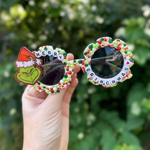 Może przedstawiać: Białe okulary przeciwsłoneczne z zielonymi, czerwonymi i białymi koralikami i kryształkami. Okulary mają napis "Merry Grinchmas" na oprawkach.