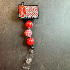 May include: A retractable badge reel with a rectangular Coca-Cola design that reads "SUSPECT RUNS ON COCA-COLA." The reel has red and pink beads and a clear plastic clip.
