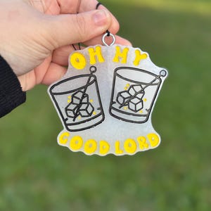 Könnte beinhalten: Weißer Glitzer-Anhänger mit schwarzer und gelber Aufschrift "Oh My Good Lord" und zwei schwarz-weißen Illustrationen von Gläsern mit Eiswürfeln.