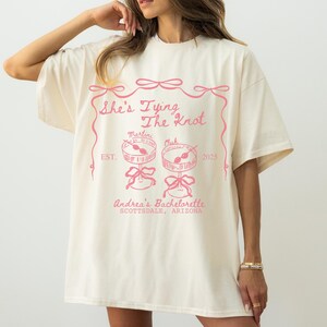 Comfort Colors® Personalized Coquette She&#39;s Tying the Knot Shirt, Custom Bow & Martini Bachelorette Party Favors, Destination Bach Tee Gift