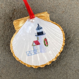 May include: A white seashell ornament with a gold rim, featuring a painted image of a lighthouse with a red roof and a wreath on the door. The ornament has a red ribbon for hanging.