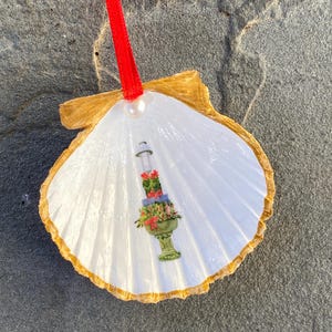 May include: A white seashell ornament with a gold-painted edge and a red ribbon. The shell features a painted floral arrangement in a green vase. A small pearl is attached to the top of the shell.