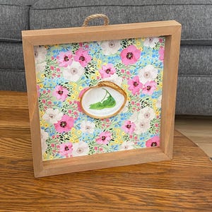 May include: A framed floral print with a small white shell with a green leaf inside. The frame is made of wood and has a light brown finish.