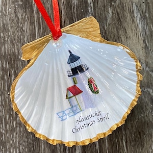 May include: A seashell Christmas ornament with a painted lighthouse scene. The white shell has a gold-painted edge and a red ribbon for hanging. The lighthouse is white with a black top and a red roof. Text reads "Nantucket Christmas Stroll."