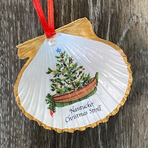 May include: A Christmas ornament made from a scallop shell, featuring a painted Christmas tree in a boat. The shell has a gold-painted edge and a red ribbon for hanging. The text "Nantucket Christmas Stroll" is printed on the shell.