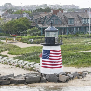 May include: A coastal scene featuring a lighthouse with an American flag design. The lighthouse is white with a black top and sits on a rocky base near a sandy beach. Houses and greenery are in the background.