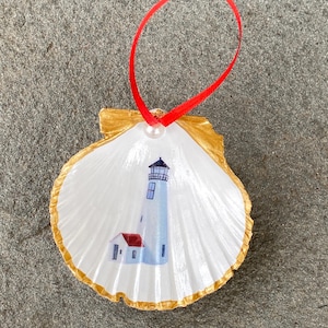 Puede incluir: Un adorno de concha blanca con ribete dorado y una imagen pintada de un faro blanco con techo rojo. El adorno tiene una cinta roja atada a la parte superior.