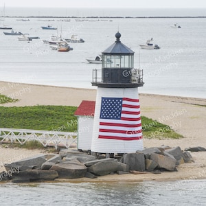May include: A white lighthouse with a black roof and a red, white, and blue American flag draped over it. The lighthouse is situated on a rocky outcropping with a sandy beach in the foreground and a body of water in the background.