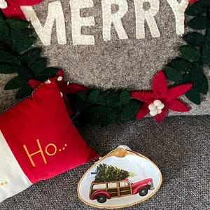 May include: A white ceramic clam shell dish with a gold rim. The dish features a painted image of a red vintage station wagon with a Christmas tree on top. The text "Ho..." is visible on a red and white pillow in the background.
