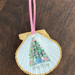 May include: A hand-painted seashell Christmas ornament. The white shell features a Christmas tree design with pink bows and gift boxes. The shell is trimmed in gold and has a pink ribbon for hanging. The text "Merry Mahjong" is written on the shell.